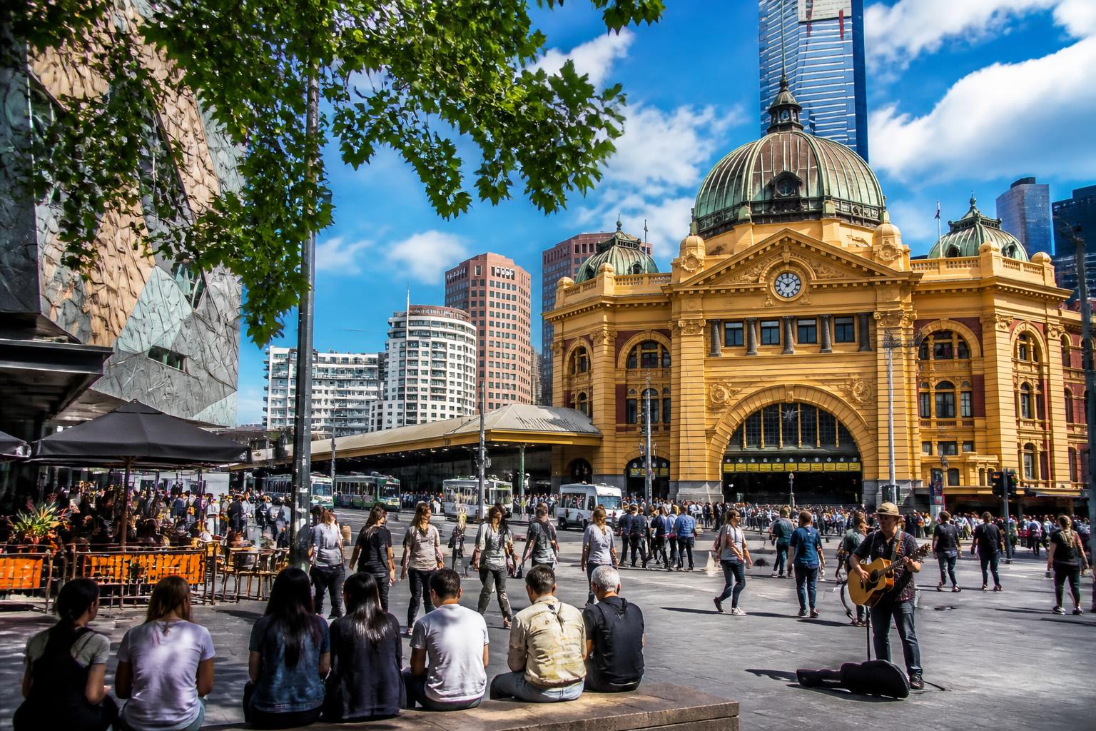 Cena movimentada de rua em Melbourne com pessoas caminhando perto da estação Flinders Street e cafés ao ar livre sob céu azul.