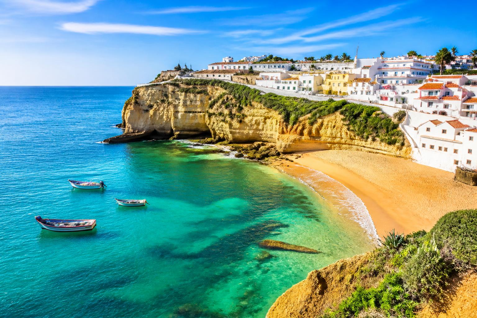 Vista da costa do Algarve com praias de areia dourada, águas turquesa, falésias rochosas e uma vila branca ao fundo.