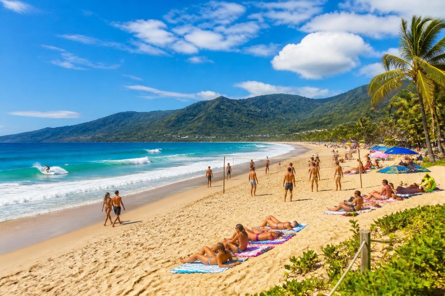 Praia de Maresias com areia clara, mar azul, pessoas surfando, tomando sol e caminhando, cercada por vegetação tropical e céu ensolarado.